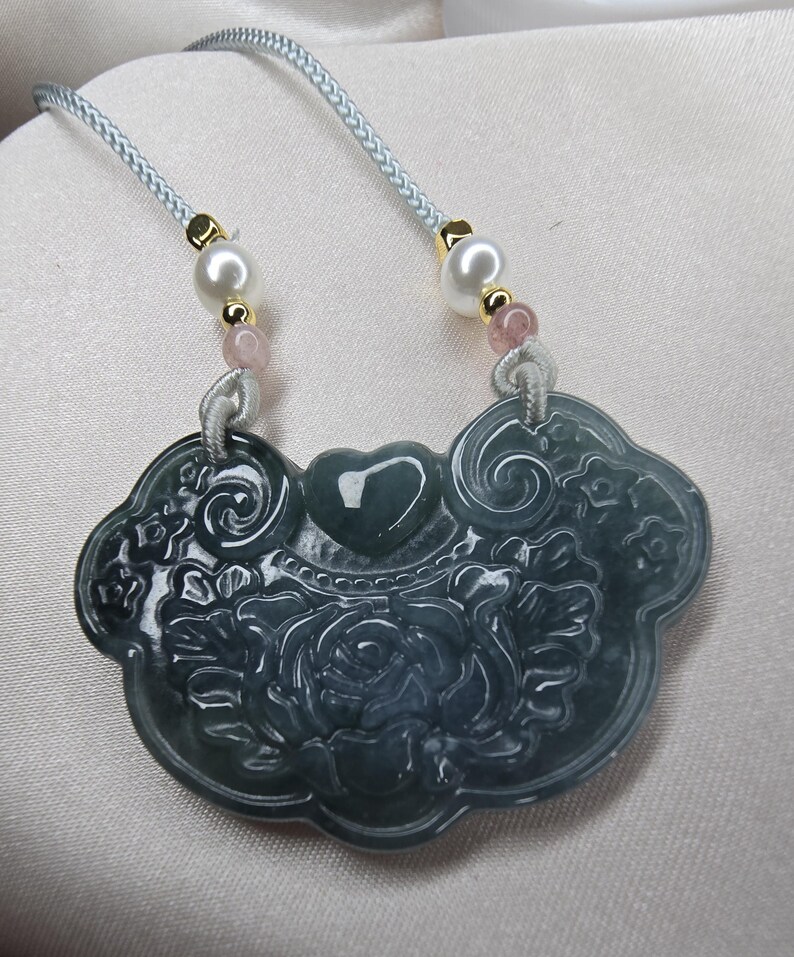 Puede incluir: Un collar con un colgante de jade tallado con un dise&ntilde;o floral y un coraz&oacute;n en el centro. El colgante est&aacute; unido a un cord&oacute;n azul claro con cuentas blancas y rosadas.