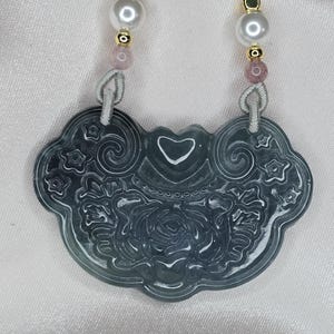 Puede incluir: Un collar con un colgante de jade tallado con un dise&ntilde;o de coraz&oacute;n y flores. El colgante es verde con detalles blancos y est&aacute; unido a un cord&oacute;n blanco con cuentas rosas y doradas.