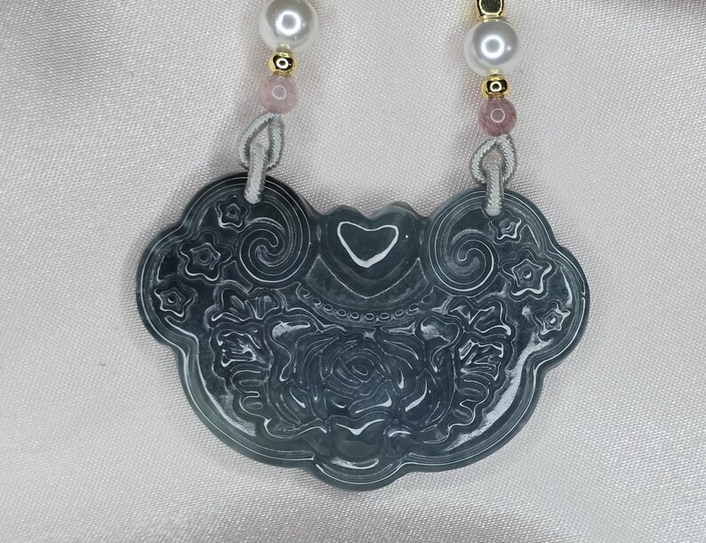 Puede incluir: Un collar con un colgante de jade tallado con un dise&ntilde;o de coraz&oacute;n y flores. El colgante es verde con detalles blancos y est&aacute; unido a un cord&oacute;n blanco con cuentas rosas y doradas.