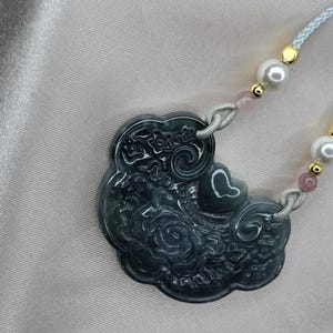 Puede incluir: Un collar con un colgante de jade verde oscuro con un dise&ntilde;o floral detallado. El colgante tiene forma de nube y cuelga de una delicada cadena de cuerda con cuentas blancas y rosadas.