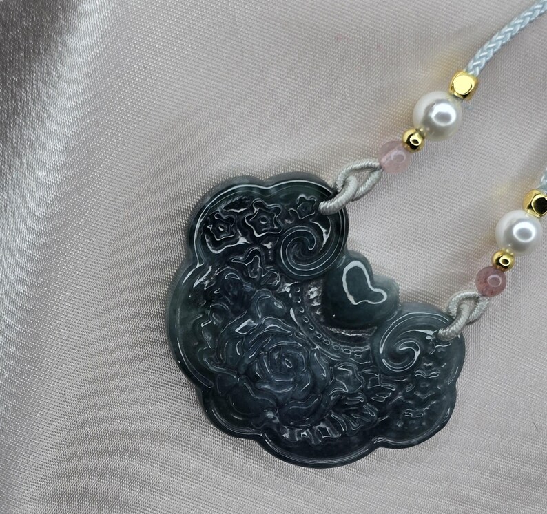 Puede incluir: Un collar con un colgante de jade verde oscuro con un dise&ntilde;o floral detallado. El colgante tiene forma de nube y cuelga de una delicada cadena de cuerda con cuentas blancas y rosadas.