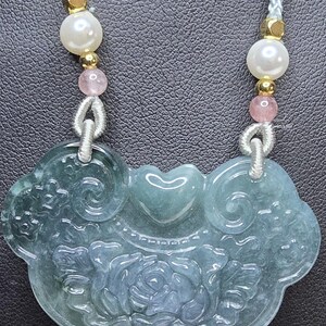 Puede incluir: Un collar con un colgante de jade tallado con un dise&ntilde;o floral. El colgante es verde y blanco y est&aacute; unido a un cord&oacute;n blanco con cuentas rosas y blancas.