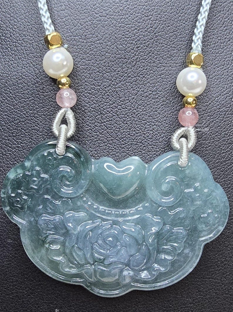 Puede incluir: Un collar con un colgante de jade tallado con un dise&ntilde;o floral. El colgante es verde y blanco y est&aacute; unido a un cord&oacute;n blanco con cuentas rosas y blancas.