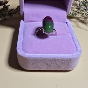 May include: A silver ring with a large, oval gemstone. The gemstone is a mix of green and red colors. The ring is presented in a purple velvet ring box.