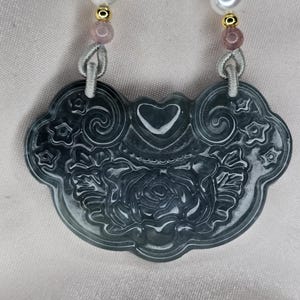 Puede incluir: Un colgante de jade verde oscuro con un dise&ntilde;o tallado de rosa y coraz&oacute;n. El colgante est&aacute; encordado con cuentas rosadas y blancas.