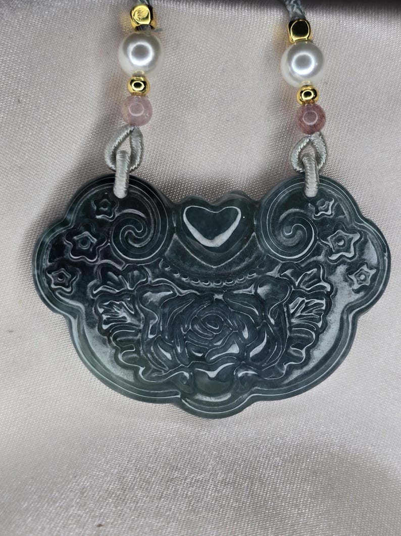 Puede incluir: Un colgante de jade verde oscuro con un dise&ntilde;o tallado de rosa y coraz&oacute;n. El colgante est&aacute; encordado con cuentas rosadas y blancas.