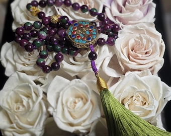 Puede incluir: Un collar de cuentas con piedras moradas y verdes, un colgante dorado decorativo y una borla verde. El colgante tiene un diseño de mosaico con detalles rojos, azules y dorados. El collar se exhibe sobre rosas blancas y rosas pálidas.