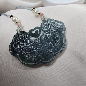 Puede incluir: Un collar con un colgante de jade tallado con un dise&ntilde;o floral. El colgante es verde y tiene un patr&oacute;n floral detallado. El collar est&aacute; hecho de un cord&oacute;n azul claro con cuentas blancas y rosadas.