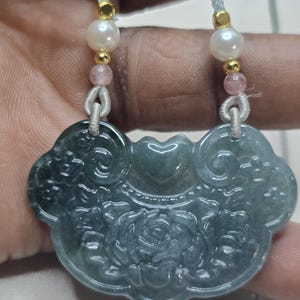 Puede incluir: Un collar con un colgante de jade tallado con un dise&ntilde;o floral. El colgante es verde con un tono azul claro y est&aacute; unido a un cord&oacute;n blanco con cuentas rosas y blancas.