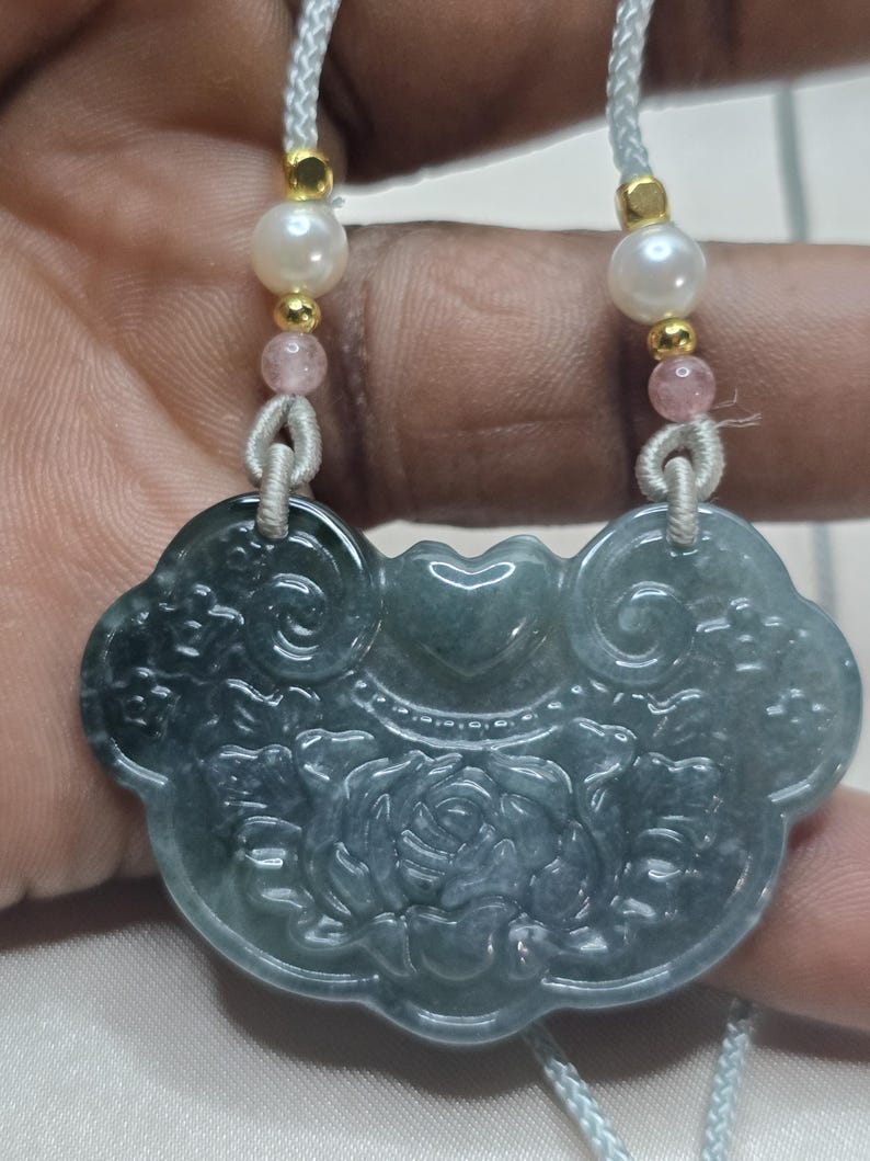 Puede incluir: Un collar con un colgante de jade tallado con un dise&ntilde;o floral. El colgante es verde con un tono azul claro y est&aacute; unido a un cord&oacute;n blanco con cuentas rosas y blancas.