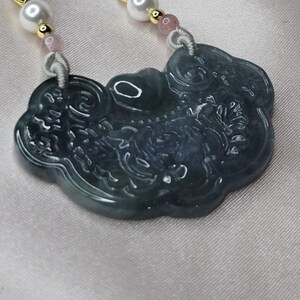 Puede incluir: Un collar con un colgante de jade tallado con un dise&ntilde;o de un drag&oacute;n y nubes. El collar est&aacute; hecho de jade azul claro y tiene cuentas de perlas blancas y rosas.
