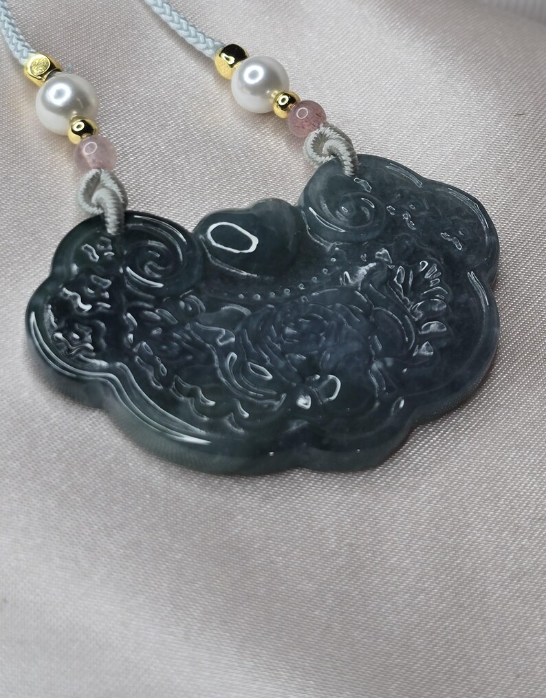 Puede incluir: Un collar con un colgante de jade tallado con un dise&ntilde;o de un drag&oacute;n y nubes. El collar est&aacute; hecho de jade azul claro y tiene cuentas de perlas blancas y rosas.
