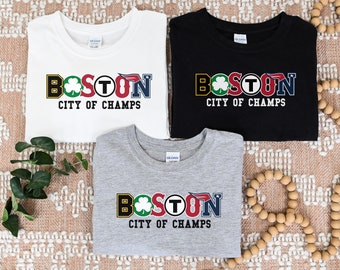 May include: Three t-shirts with the word "BOSTON" in colorful letters and the text "CITY OF CHAMPS" below. The shirts are white, black, and gray. The word "BOSTON" incorporates sports team logos.
