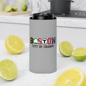 May include: A gray can cooler with the word "BOSTON" in colorful letters and the text "CITY OF CHAMPS". The cooler is designed to hold a standard-sized can and keep beverages cold. The background includes cut lemons and limes.