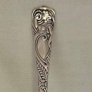 May include: A silver spoon with an ornate handle featuring a detailed floral and figure design. The spoon's bowl is oval-shaped, and the handle has a slender, elongated form. The spoon is likely made of sterling silver.