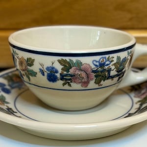 May include: A white teacup and saucer with a blue and floral design. The cup has a blue band around the rim and a floral pattern with pink, blue, and green accents. The saucer has a matching floral design.