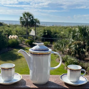 May include: A white porcelain tea set with gold and navy blue trim, including a teapot, two teacups, and saucers. The set is displayed on a wooden surface with a scenic background of greenery and a view of the ocean.
