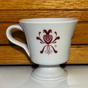 May include: White ceramic teacup with a red stylized heart design and a bird motif. The cup has a handle and a pedestal base.