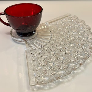 May include: A deep red glass teacup resting on a clear glass snack plate. The plate is shaped like a fan with a textured, starburst-like pattern across its surface and a smooth, circular indentation designed to hold the base of the teacup securely.
