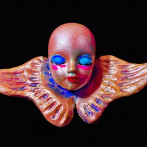 May include: A ceramic angel figurine with wings, a pink face, blue eyes, and colorful paint details. The angel has a ruffled collar and is painted in a metallic finish.