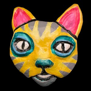 Könnte beinhalten: Eine handbemalte Katzenmaske mit leuchtenden Farben. Die Maske hat eine gelbe Basis mit grauen Streifen und Akzenten, blaue und türkisfarbene Augenränder, rosa Innenohren und weiße Augen. Die Katzennase ist türkis und der Mund grau.