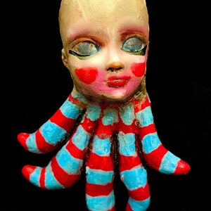 Puede incluir: Una escultura caprichosa pintada a mano de una cabeza de muñeca con tez pálida, mejillas rojas y labios rojos. Los brazos de la muñeca están diseñados como tentáculos a rayas en rojo y azul claro alternados. El fondo es negro sólido.