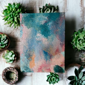 Puede incluir: Una pintura cuadrada sobre lienzo con pinceladas abstractas en tonos azules, verdes, rosas y naranjas. La pintura está rodeada de pequeñas suculentas en macetas.