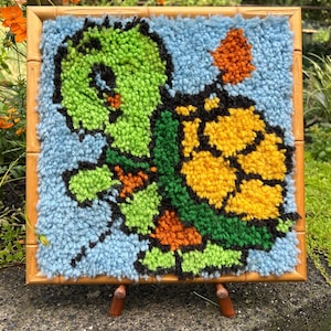 以下が含まれることがあります： 手作りのカラフルなカメの壁掛け。カメは、水色の背景に緑、黄色、オレンジの色合いで描かれています。このアートワークは天然木のフレームに収められ、小さな木製のイーゼルに飾られています。