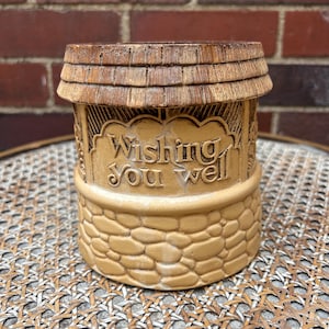 May include: A tan ceramic wishing well with a wooden shingle roof. The well features the words "Wishing You Well". The base is made of textured stones. A decorative item.