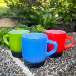 Puede incluir: Tres tazas coloridas en verde neón, azul y rojo, cada una con una base negra. Las tazas tienen una forma redondeada con un asa. Las tazas están sobre una superficie de piedra, con vegetación en el fondo.