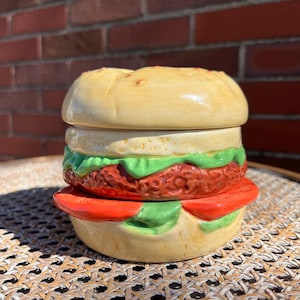 Puede incluir: Un recipiente de cerámica con forma de hamburguesa, con pan, lechuga, una hamburguesa y una rodaja de tomate. El recipiente está pintado con colores y detalles realistas, y probablemente se utiliza para guardar cosas o decorar.