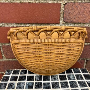 Peut inclure: Un panier mural en demi-cercle de couleur beige avec un motif tissé. Le panier a un dessus décoratif avec des détails arqués et de petites poignées latérales. Le panier est fait d'un matériau brun clair et est monté sur un mur de briques rouges.