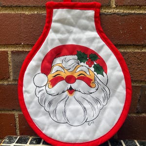 Puede incluir: Un guante de cocina navideño blanco con borde y lazo rojos. El guante presenta una cara sonriente de Papá Noel con un gorro rojo, mejillas rosadas y barba blanca. Acebo y bayas están encima de la cabeza de Papá Noel.