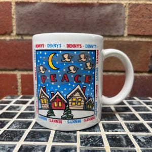 May include: White ceramic mug with a winter scene featuring the word "PEACE" and the text "DENNY'S" around the rim. The design includes houses, a crescent moon, and flying birds against a blue background.