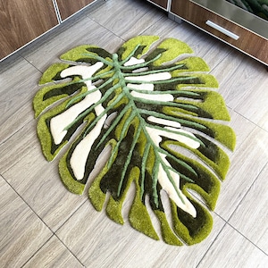 Può includere: Un tappeto a forma di foglia nei toni del verde, bianco e verde scuro. Il tappeto ha un aspetto strutturato e trapuntato, con un design dettagliato che imita le venature e la forma di una foglia di Monstera. Il tappeto è posizionato su un pavimento piastrellato.
