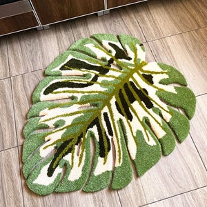 Puede incluir: Una alfombra verde y blanca con forma de hoja tropical grande. La alfombra tiene una superficie texturizada y esponjosa.