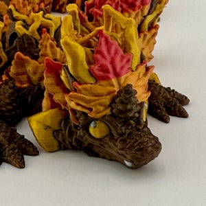 Könnte beinhalten: Eine 3D-gedruckte Figur eines Drachen mit braunem Körper und gelben, orangefarbenen und roten Blättern, die seinen Rücken und Kopf bedecken.