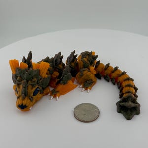 May include: A small, orange and brown dragon figurine with a textured surface. The dragon is curled up with its tail wrapped around its body. The figurine is about the size of a quarter.