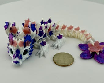 May include: A white, articulated dragon figurine with blue, purple, and pink accents. The dragon is next to a purple and pink flower-shaped object. A coin is next to the dragon.