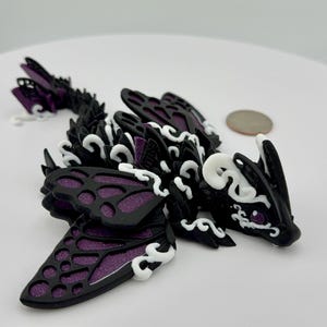 May include: A black and purple 3D printed dragon figurine with white accents. The dragon has butterfly wings and a curled tail.
