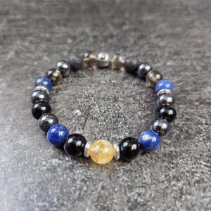 Puede incluir: Un brazalete con cuentas negras, azules y doradas. El brazalete tiene un dije de plata.