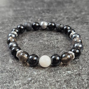 Puede incluir: Un brazalete de cuentas negras con detalles plateados y un dije de piedra blanca. El brazalete presenta cuentas de ónix negro, roca volcánica, cuarzo ahumado y hematita.