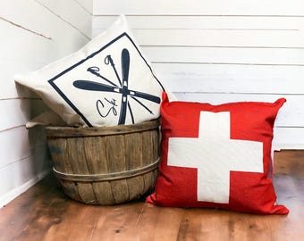 May include: Two decorative throw pillows. One is cream-colored with a black ski graphic and the word "Ski". The other is red with a white cross. They are next to a wooden basket.