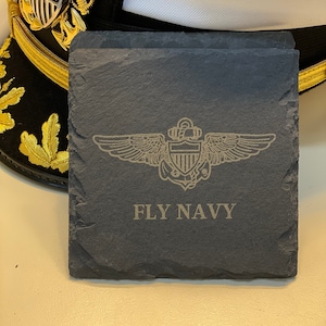 May include: Dark gray slate coasters with the words "FLY NAVY" and a detailed emblem. A navy officer's hat with gold trim and a silver emblem is in the background.