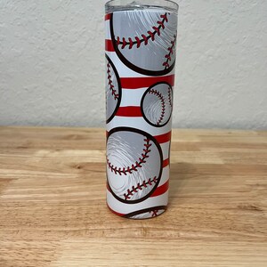 Pode incluir: Um copo de aço inoxidável branco e vermelho com um padrão de beisebol. O copo tem listras vermelhas e bolas de beisebol brancas com costuras vermelhas.
