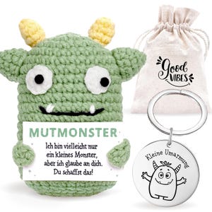 Könnte beinhalten: Ein grünes gehäkeltes Monstertier mit gelben Akzenten, das ein Schild mit der Aufschrift "MUTMONSTER" hält. Enthält einen Kordelzugbeutel mit "Good Vibes" und einen Schlüsselanhänger mit einer Monstergrafik und den Worten "Kleine Umarmung".