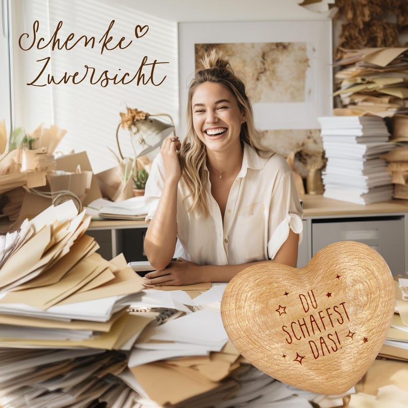 May include: A wooden heart-shaped ornament with the text "DU SCHAFFST DAS!" in red lettering. The ornament is surrounded by papers and a woman is smiling in the background.