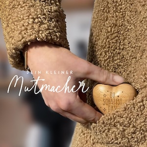 May include: A wooden heart-shaped keychain with the text "DU SCHAFFST DAS" engraved on it. The keychain is being held in a person's hand, with the text "DEIN KLEINER MUTMACHER" visible in the background.