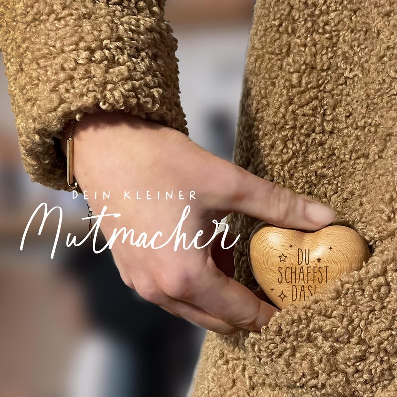 May include: A wooden heart-shaped keychain with the text "DU SCHAFFST DAS" engraved on it. The keychain is being held in a person's hand, with the text "DEIN KLEINER MUTMACHER" visible in the background.