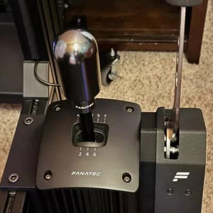 Puede incluir: Un cambio de marchas de carreras negro con una perilla plateada y una palanca de freno de mano negra. El cambio de marchas tiene un patrón de marchas con los números 1, 2, 4, 5, 6 y 7, así como la letra R para la marcha atrás. El cambio de marchas también tiene la palabra "FANATEC" impresa en él.